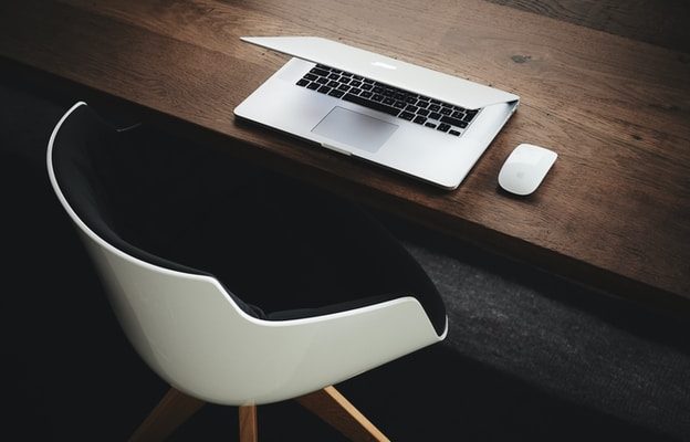Half dichtgeklapte laptop, met muis ernaast op een houten bureau met daarvoor een witte stoel